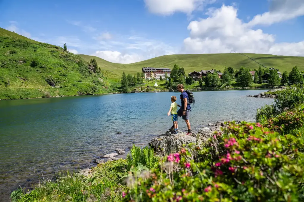 See in den Nockbergen mit klarem Wasser, umgeben von grünen Almwiesen und sanften Hügeln; im Vordergrund stehen ein Erwachsener und ein Kind auf einem Felsen am Ufer und blicken über die Landschaft.